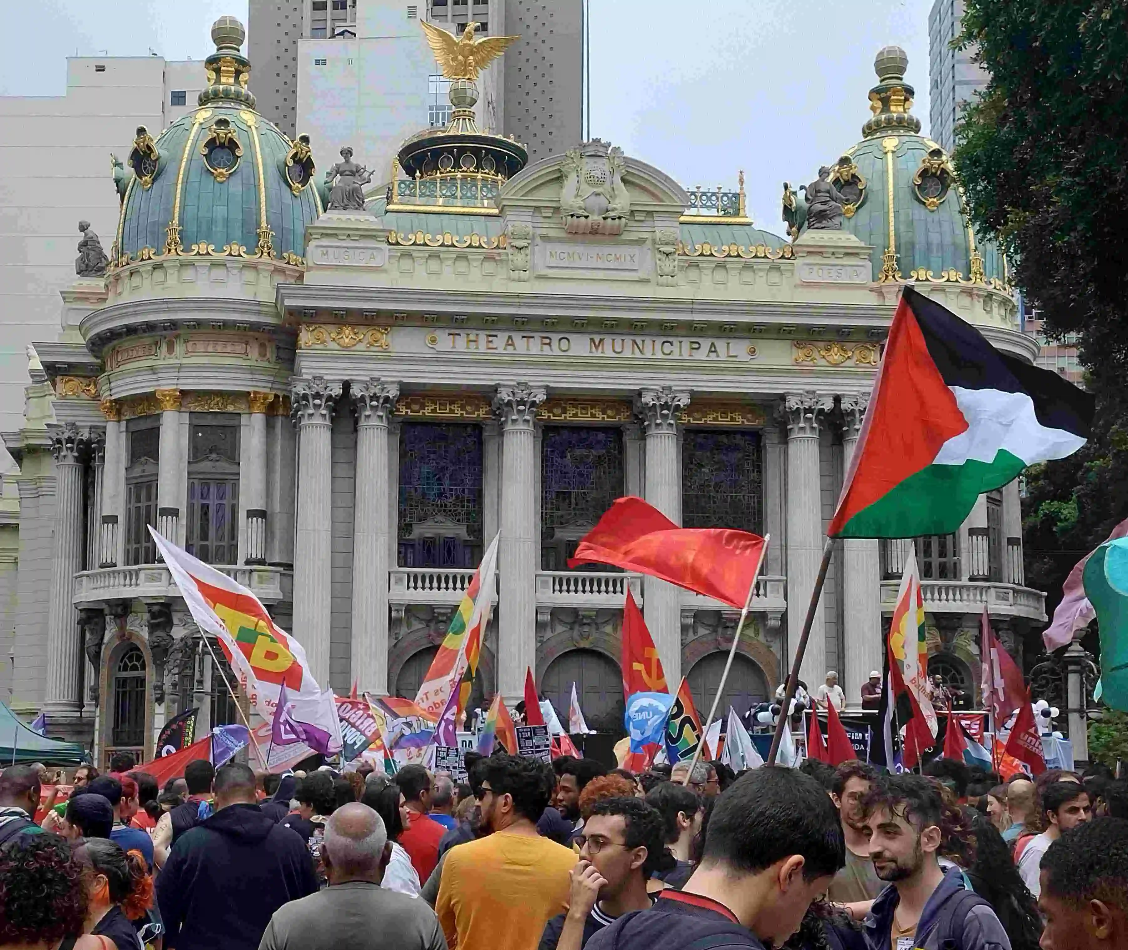 multidão (apenas as cabeças visíveis na foto) segurando bandeiras em frente ao theatro municipal do rio de janeiro. em destaque: uma bandeira da palestina, algumas da ctb (central dos trabalhadores do brasil) e outras bandeiras vermelhas