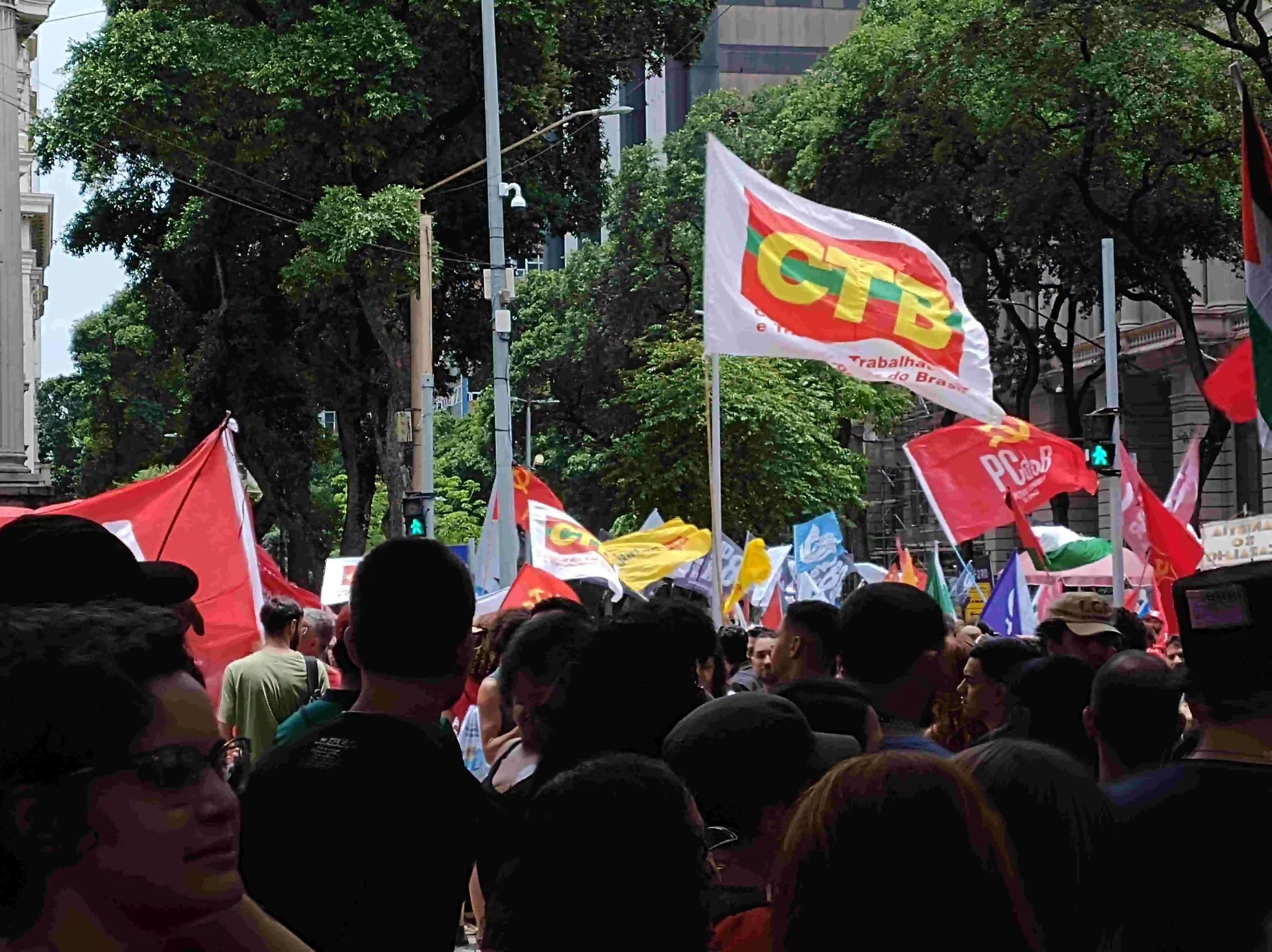 foto de dentro da multidão para fora na avenida, destacando uma bandeira do ctb (central de trabalhadores do brasil) e uma do pcdob ao lado. prédios e árvores ao fundo